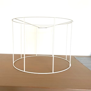 May include: A white metal lampshade frame. The frame is cylindrical, with a circular top and base, and vertical supports. The frame sits on a brown wooden surface against a white background. The frame is empty.