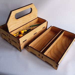 Puede incluir: Una caja de herramientas de madera con asa y múltiples compartimentos. La caja es de madera clara con bordes marrones oscuros. Varios destornilladores con mangos amarillos y negros son visibles dentro del compartimento superior abierto.