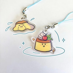 May include: Two clear acrylic charms with light blue lanyards. One charm is a yellow and brown pudding with a sad face. The other charm is a smiling pudding with whipped cream, a cherry, and green leaves.