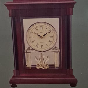 May include: A wooden mantel clock with a gold face and Roman numerals. The clock has a rotating pendulum and the words "Mantel Clock with Rotating Pendulum" and "Stiefel" are printed on the box.