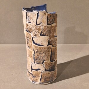 Puede incluir: Un jarrón cilíndrico de cerámica con una superficie texturizada que presenta un patrón de mosaico de formas rectangulares en tonos bronceado y marrón, separadas por líneas de unión azul oscuro. El borde interior tiene un esmalte azul liso.