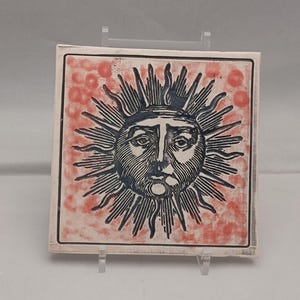 May include: Square ceramic tile featuring a detailed sun design with a face. The sun is depicted in dark blue against a red background with a speckled pattern. The tile has a white border and is displayed on a clear stand.