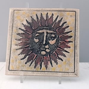 May include: A square ceramic tile featuring a central embossed sun face with radiating rays in black and muted red tones. The background is a textured off-white surface decorated with scattered yellow dots, all enclosed within a thin black border.