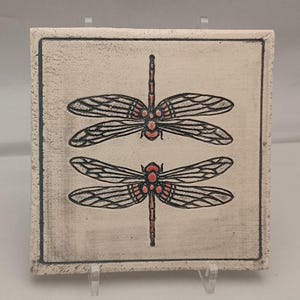 May include: Square ceramic tile featuring two dragonfly illustrations. The dragonflies have black wings with detailed veining and red bodies. The tile has a cream-colored background with a dark border.