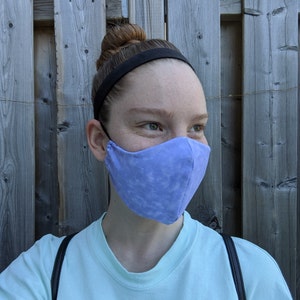 May include: A person wearing a light blue face mask with a white tie-dye pattern. The mask is secured with black elastic ear loops. The person is wearing a light blue shirt and has a black headband on their head.