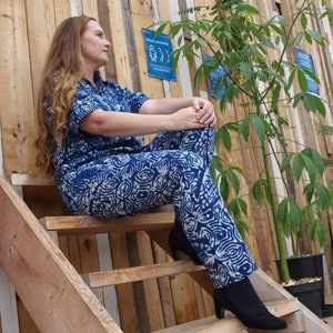 Op de afbeelding: Een vrouw in een blauwe en witte gebloemde jumpsuit zit op houten treden. De jumpsuit heeft een losse pasvorm en een kraag. De vrouw draagt zwarte hakken.