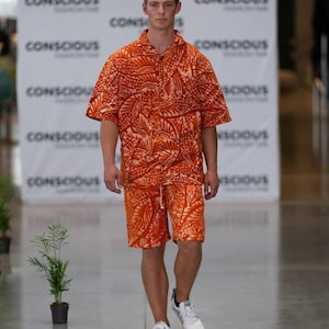 Peut inclure: Un mannequin défile sur un podium portant une chemise à manches courtes à motifs orange et blanc et un short assorti. La chemise a un devant boutonné et le short a une taille élastique. Le mannequin porte des baskets blanches.