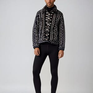 Op de afbeelding: Een zwart-wit gepatroneerd jack met een mock neck shirt en zwarte leggings. De persoon draagt zwarte schoenen.