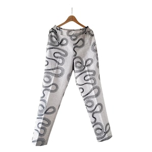 May include: A pair of light-colored pants with a black, dotted, swirling pattern. The pants are hanging on a wooden hanger against a white background. The pattern resembles a series of connected, abstract shapes.