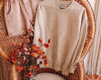 May include: A beige sweatshirt and pink shorts are displayed on a wicker chair. A vase of dried orange flowers sits below the clothing. The sweatshirt is hanging on a white hanger.