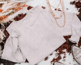 May include: A light beige sweatshirt with ruffled sleeves and a matching cropped top are displayed on a cowhide rug. A layered gold necklace and a pair of embroidered cowboy boots complete the ensemble. Dried floral accents add a touch of rustic charm.