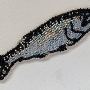 May include: A beaded fish-shaped applique. The fish features a light blue body with black and silver beaded details. Accents of light blue beads are scattered throughout the design. The applique is outlined with white beads.