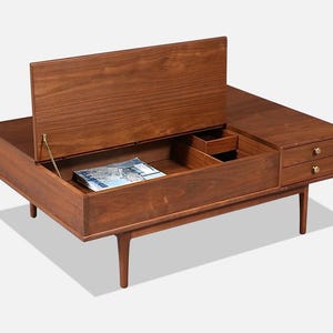 May include: A mid-century modern coffee table made of wood, with a lift-top compartment and two drawers. The table has a rich brown finish, brass hardware, and tapered legs. A magazine is visible inside the open compartment.