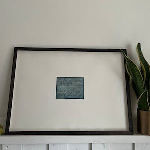 Puede incluir: Una impresión abstracta en blanco y negro con un tinte azul enmarcada. La impresión está en un marco negro y está colgada en una pared blanca. Una planta verde está en una maceta dorada en el estante debajo de la impresión.