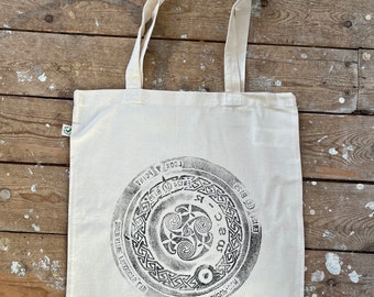 May include: A white canvas tote bag with a black circular design featuring a Celtic knot and the text "Look at the bright side of life, humour club".