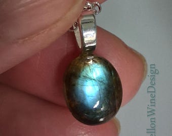May include: An oval-shaped labradorite pendant featuring a polished, dark brown surface with a vibrant, iridescent blue flash in the center. The gemstone is set in a smooth, silver-toned metal bail attached to a chain. The text MellonWineDesign is visible.