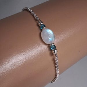 May include: A silver chain bracelet featuring a central oval moonstone bead flanked by two small metallic silver beads and two translucent blue glass beads. The design is symmetrical, with the beads centered on a delicate silver-toned link chain.