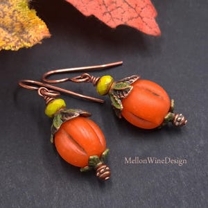 May include: A pair of dangle earrings featuring orange pumpkin-shaped beads with green metal leaf caps, small yellow accent beads, and copper-toned ear wires. The text MellonWineDesign is visible in the lower right corner of the image.