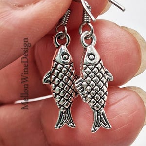 May include: A pair of silver-colored fish earrings. Each earring features a detailed fish pendant with scales and fins, suspended from a hook. The earrings are held in a person's hand, with the text "Mellon Wine Design" visible.