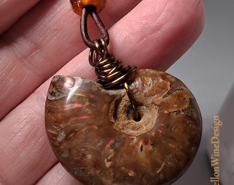 May include: A brown ammonite fossil pendant with copper wire detailing and a leather cord. The fossil displays iridescent colours and an orange bead. A unique piece of jewellery.