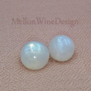 May include: Two round, white, iridescent cabochon stones with a smooth, polished surface. The text MellonWineDesign is visible in the upper portion of the frame. These circular, dome-shaped items exhibit a soft luster and reflective quality.