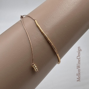 May include: A delicate bracelet featuring a single row of small, metallic gold-colored cylindrical beads strung on a thin, light brown cord. The adjustable cord ends with three matching gold-colored beads. The text Mellon WineDesign is visible.