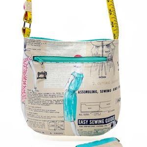 May include: A cream-colored crossbody bag with a sewing pattern print, a teal zipper, and a yellow tape measure strap. A matching small pouch is also present. The bag features a sewing machine charm and the text "EASY SEWING GUIDE".