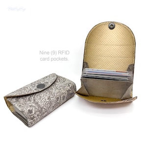May include: A silver and gray RFID card holder with a floral pattern, open to reveal nine card pockets. The interior is a light tan color. The text "Nine (9) RFID card pockets" is visible.