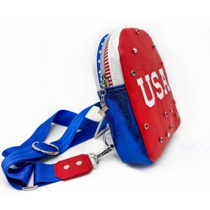 May include: A red, white, and blue crossbody bag with a zipper closure and a star-shaped zipper pull. The bag has a blue metallic strap and features the word "USA" in white letters on the front. The bag is decorated with rhinestones.