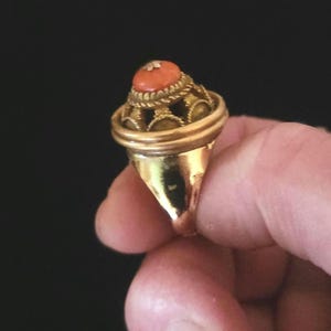 May include: A gold ring featuring a prominent, rounded orange stone set atop a decorative, openwork cage structure. The metal band is smooth and tapers toward the bottom, while the setting includes intricate twisted wire detailing around the base.