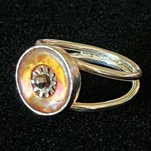 May include: A silver-toned ring featuring a circular setting with an iridescent orange and yellow center. A small, metallic, gear-shaped component with a dark, faceted bead sits in the middle of the circular face. The band is split into two.
