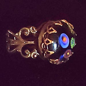 May include: A ring featuring a dark, rounded glass cabochon with embedded colourful floral-like patterns in blue, orange, and green. The setting is made of an ornate, metallic filigree band with intricate scrollwork and a decorative metal crown.