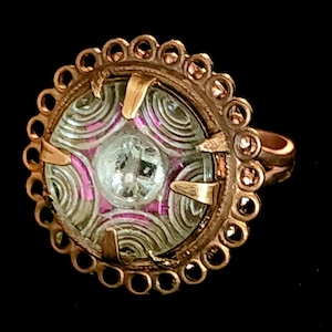 May include: A copper ring with a circular design and a clear glass stone with pink and white swirls. The stone is set in a bezel setting and the ring is decorated with a scalloped edge.