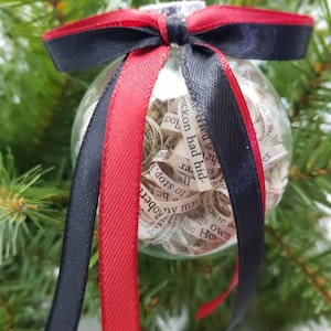 Puede incluir: Un adorno navideño de vidrio transparente con un lazo de cinta roja y negra. El adorno está lleno de trozos de papel enrollados con texto impreso en ellos. El texto incluye las palabras "the How", "is own", "uckon had hid", "1001", "de", "Arc", "ra", "all", "8", "peyman", "to stop", "he San", "Robert", "dis", "er", "sons", "ns".