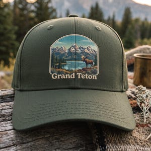 Op de afbeelding: Een olijfgroene baseballpet met een grafische print van een bergketen, een meer en een eland. De tekst "Grand Teton" is in een crèmekleurig lettertype onder de illustratie gedrukt. De pet heeft een gestructureerde kroon en een gebogen klep.