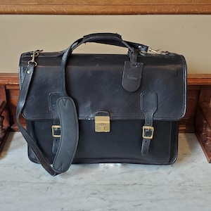 May include: A black leather briefcase with a gold buckle and a shoulder strap. The briefcase has a label that says "Bridgman".
