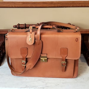May include: A brown leather briefcase with a long strap and a brass lock. The briefcase has two buckles on the front and a small leather tag with the letter "H" on it.