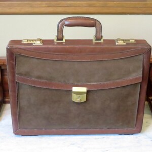 May include: A brown leather briefcase with a suede exterior and a brass lock. The briefcase has a top handle and two side clasps.