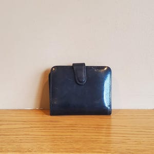 May include: A small, dark blue leather wallet with a glossy finish. The wallet has a rectangular shape with rounded corners and a snap closure. It is sitting on a wooden surface.