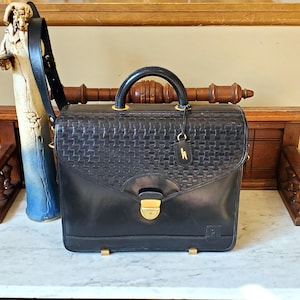 May include: Black leather briefcase with a woven pattern and a gold clasp. The briefcase has a shoulder strap and a small gold tag.