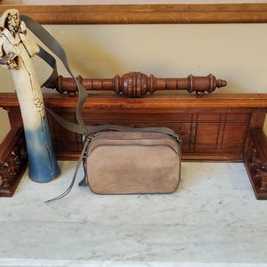 May include: A tan leather crossbody bag with a long strap rests on a marble surface. A ceramic figure of a man in a long coat and a wooden decorative piece are behind the bag. The bag has a rectangular shape.