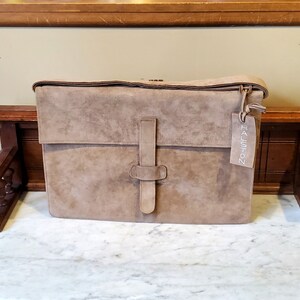 May include: A large, brown suede leather briefcase with a Halston tag attached. The bag has a flap closure with a buckle and a strap.