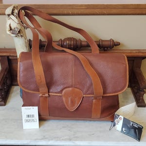 May include: A brown leather handbag with tan straps and a flap closure. The bag features a textured surface and a small emblem on the front. A tag with text is attached to the bag.