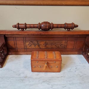 May include: A rectangular wooden wall shelf with a carved back panel and a decorative spindle top rail, featuring side supports. In front sits a small, tan leather-covered box with metal stud detailing, a hinged lid, and a leather tassel pull.