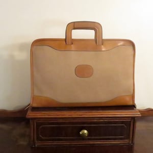 May include: A tan leather briefcase with a brown leather handle and a brown leather patch on the front. The briefcase is sitting on a wooden surface.