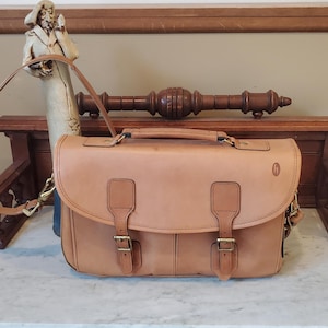 May include: A tan leather satchel featuring a top handle, an adjustable shoulder strap, and a front flap secured by two buckle straps. The bag includes a small embossed logo on the right side and antique brass-toned metal hardware throughout.