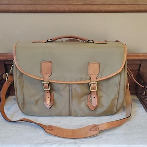 May include: A tan nylon messenger bag featuring a top handle and an adjustable shoulder strap. The bag has a large front flap secured by two tan leather straps with brass buckles. The edges of the flap and the strap attachments are trimmed in leather.