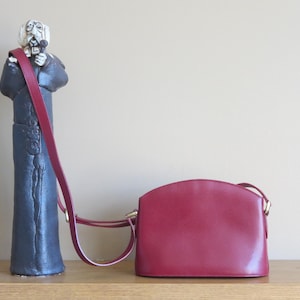 May include: A burgundy leather crossbody bag with a curved top and gold-tone hardware. A tall, dark statue with a red strap is to the left of the bag. The bag is on a wooden surface.