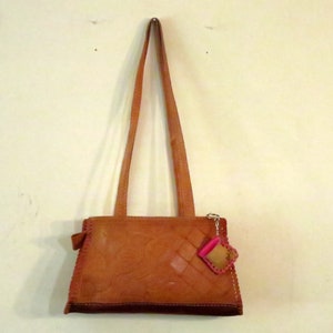 以下が含まれることがあります： ピンクとブラウンのチャームが付いたブラウンの革製のショルダーバッグ。バッグは織り模様で、フラップで閉じます。