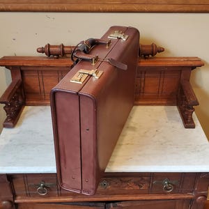 May include: A brown leather briefcase with gold-colored combination locks and hardware. The briefcase has a handle and a leather tag. It is resting on a marble-topped wooden table with decorative carvings and drawers.